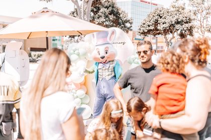 A costumed bunny mascot in a top hat and plaid vest greeting families beside a pastel balloon arch at a Dreamers Markets pop-up